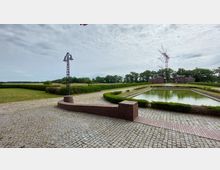 Weitläufige Freifläche mit einem rechteckigen, von Hecken umgebenen Wasserbecken in der Mitte, gepflastertem Boden und einer kleinen Backsteinmauer im Vordergrund. Im Hintergrund stehen flache Backsteingebäude, Bäume und ein großer Sendeturm.