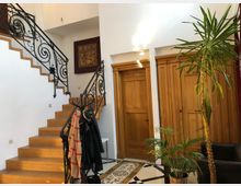 Eingangsbereich eines Wohnhauses mit einer Holztreppe und kunstvoll verziertem Metallgeländer. Im Vordergrund stehen eine Topfpflanze und eine Garderobe, im Hintergrund sind Holztüren und ein farbiges Wandbild zu sehen.