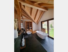 Modernes Badezimmer mit freistehender Badewanne und separatem Waschbecken, Holzdetails an der Decke und den Möbeln sowie großen Fenstern mit Blick auf einen Garten. Schwarze Fliesen und natürliche Materialien dominieren das Design.