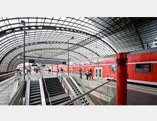 Ein moderner Bahnhof mit gläsernem, geschwungenem Dach, darunter Gleise, Bahnsteige und ein roter Regionalzug. Im Vordergrund führen Rolltreppen und Treppen auf die obere Ebene, während Passagiere auf den Bahnsteigen zu sehen sind.