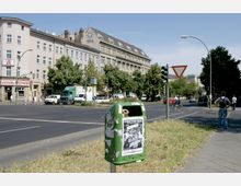 Straßenszene mit Kreuzung, moderner mehrstöckiger Wohn- und Geschäftsgebäudezeile im Hintergrund, darunter eine Apotheke sowie Eingänge zur U-Bahn. Ein grüner Mülleimer mit Plakaten steht im Vordergrund, umgeben von Fußwegen, Autos und Bäumen.