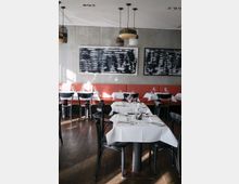 Ein modernes Restaurant mit dunklem Holzboden, schwarzen Stühlen und weiß gedeckten Tischen. An der Wand hängen abstrakte Kunstwerke in Schwarz-Weiß, darüber hängen mehrfarbige, minimalistische Pendelleuchten.