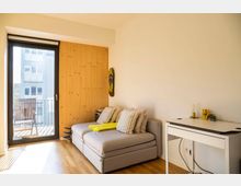 Wohnzimmer mit hellem Holzboden, einer grauen Couch mit Kissen, einem weißen Schreibtisch mit Dekoration und einer großen Glastür, die zu einem Balkon mit Stuhl führt. Eine mit Holzpaneelen verkleidete Wand befindet sich neben der Couch.