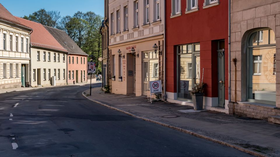 Curving paved street in a small town lined with two-story historic buildings in light stucco and brick, featuring arched doorways and shopfront windows. A sidewalk with street lamps and a “30 zone” traffic sign runs along the buildings, with trees visible at the far end of the road.
