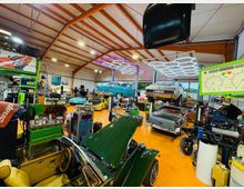 Große Autowerkstatt mit hoher Hallendecke und orangefarbenem Boden, voll mit Werkzeugwagen, Regalen und Ersatzteilen. Mehrere Oldtimer stehen auf Hebebühnen, darüber hängen helle, sechseckige LED-Leuchten und eine große Anzeigeinstrument-Tafel ist im Vordergrund zu sehen.