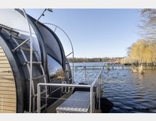 Schwimmendes Ferienhaus an einem Steg auf einem See, mit gebogener Metall- und Holzkonstruktion sowie Leiter. Im Hintergrund befinden sich weitere Stege, Schilf und Bäume bei klarem Wetter.