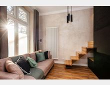 Modernes Wohnzimmer mit großem bodentiefem Fenster, grauen Vorhängen und Ecksofa mit Kissen auf Holzfußboden. An der Wand hängen ein hoher weißer Heizkörper und schwarze Pendelleuchten; rechts führt eine schwebende Holztreppe an einer dunklen Wand nach oben.