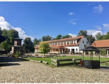 Hof mit gepflasterten Wegen, grünen Rasenflächen und einem großen Fachwerkhaus mit rotem Ziegeldach und weißen Wänden im Hintergrund. Links ein kleiner gemauerter Turm, umgeben von Bäumen und weiteren Gebäuden.