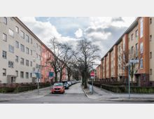 Straßenansicht mit mehrstöckigen Wohngebäuden aus der Nachkriegszeit in hellen Farben (Beige und Orange) auf beiden Seiten. In der Mitte verläuft eine schmale Pflasterstraße mit geparkten Autos, gesäumt von Bäumen ohne Blätter.