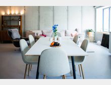 Heller, moderner Ess- und Wohnbereich mit großem weißen Esstisch und gepolsterten Stühlen, dahinter ein Sofa vor einer Sichtbetonwand. Rechts sorgen große Fenster für Tageslicht; auf dem Tisch stehen Blumen und eine Obstschale, links ist ein Regal aus Holz zu sehen.