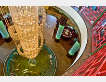Rundes Atrium in einem asiatisch inspirierten Gebäude mit einem großen Kronleuchter aus goldenen Ketten, der über einem Teich mit Koi-Fischen hängt. Um das Atrium befinden sich verzierte rote Balustraden, grüne beleuchtete Säulen und runde Esstische.