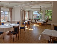 Heller Gastraum mit großen Fenstern, durch die Tageslicht einfällt, ausgestattet mit Holzboden, blauen Polsterstühlen und weiß eingedeckten Tischen. Ein Kronleuchter hängt in der Mitte, und die Fenster bieten einen Blick ins Freie mit Bäumen und einem Gebäude.