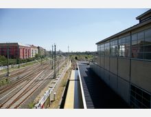 Blick auf mehrere Bahngleise und Bahnsteige in einer städtischen Umgebung, mit einem modernen Gebäude mit großen Fensterfronten rechts und mehrstöckigen Wohnhäusern links. Im Hintergrund ist der Berliner Fernsehturm sichtbar.