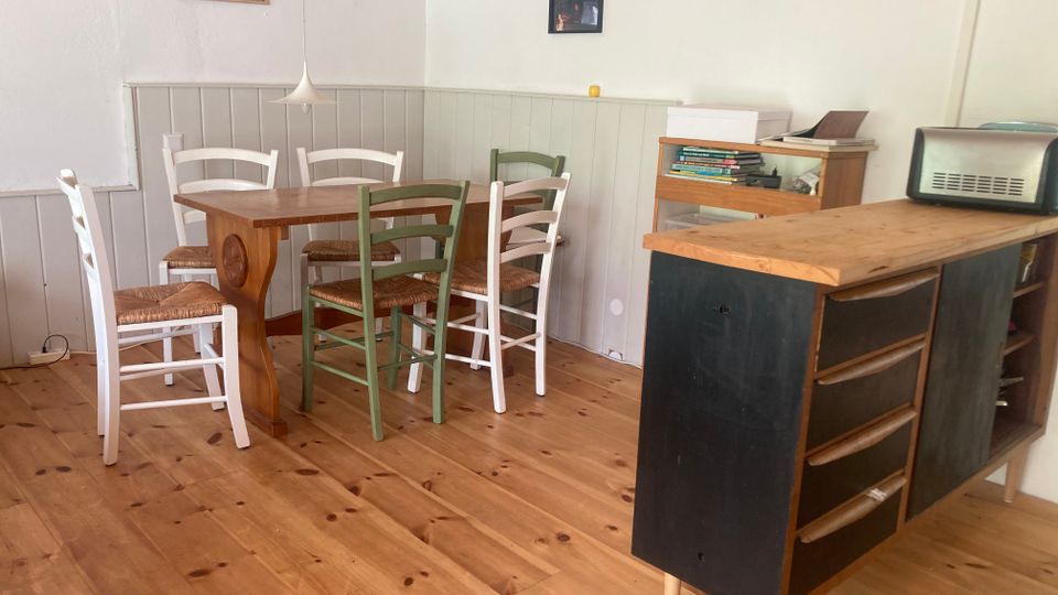 Heller Essbereich mit Holzdielenboden, weiß vertäfelten Wänden und einem rechteckigen Holztisch, umgeben von gemischten Holzstühlen. Rechts steht ein Küchenblock mit Holzarbeitsplatte und Schubladen, daneben ein kleines Regal; an der Wand hängen zwei Bilder und eine Pendelleuchte.