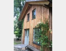 Holzhaus mit vertikaler Holzverkleidung und großen, sprossenverglasten Fenstern. Vor dem Gebäude stehen eine Holzbank und ein runder Holztisch, während Kletterpflanzen und Bäume die Umgebung prägen.