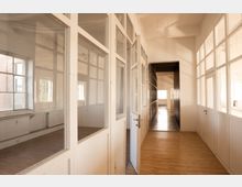 Langer Flur in einem hellen Altbau- oder Industriegebäude mit Holzfußboden und hohen Decken. Beidseitig stehen weiße, verglaste Trennwände und Türen mit großen Fensterfeldern, durch die Tageslicht in die angrenzenden Räume fällt.