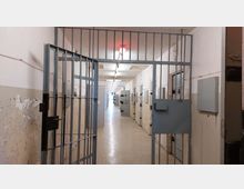 Langer Flur in einem Gefängnis mit Zellenreihen auf beiden Seiten, abgeschlossenen Türen und einer offenen Metallgittertür im Vordergrund. Die Wände sind teilweise abgeblättert, und die Decke ist mit Leuchtstofflampen ausgestattet.