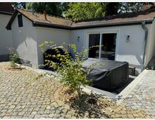 Kleiner Innenhof mit Kopfsteinpflaster und einem weißen eingeschossigen Gebäude mit Schiebetür und Satteldach. Im Vordergrund befinden sich zwei kleine, in Kies gefasste Pflanzen sowie ein mit einer schwarzen Plane abgedecktes Möbelstück.