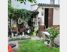 Ein kleiner Garten mit einer Rasenfläche und einem weißen Schuppen mit Holztür und schrägem Dach. Links eine überdachte Ecke mit Rankpflanzen und dekorativen Gegenständen wie Gießkannen, einem Strohhut und Laternen, sowie ein schmiedeeisernes Geländer.