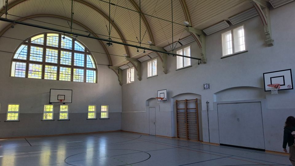 Innenansicht einer Sporthalle mit hohem Tonnengewölbe, großen Rundbogenfenstern und mehreren Basketballkörben an den Wänden. Der Hallenboden ist mit Spielfeldlinien markiert; an einer Seitenwand sind Sprossenwand und Türen zu sehen.
