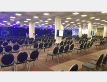 Ein großer, moderner Konferenzraum mit zahlreichen Stuhlreihen, die auf eine Bühne mit Vorhängen und blauer Beleuchtung ausgerichtet sind. Weiße Säulen und mehrere Bildschirme, die Informationen anzeigen, strukturieren den Raum.