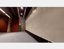 Innenraum einer Bühne mit seitlichem Blick auf eine große Leinwand. Links befinden sich rote, geriffelte Wandverkleidungen und ein Vorhang, während im Hintergrund eine Tür und technische Einrichtungen sichtbar sind.