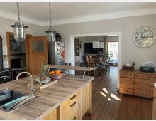 Küche mit Holzarbeitsplatte, Spüle und Hängelampen im Vordergrund. Im Hintergrund ein Essbereich mit Holzboden, Esstisch, Stühlen und Zugang zu einer Terrasse, sowie eine große Wanduhr und Kommoden.