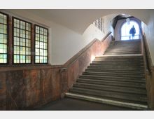 Treppenhaus mit steinerner Treppe und braunen Wandverkleidungen, seitlich Fenster mit Sprossen und buntem Glas. Im oberen Bereich führt die Treppe zu einem großen, hellen Rundbogenfenster mit einer silhouettierten Person.