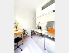 Kleine Küche mit schlichtem Design, bestehend aus einem Tisch mit Holzplatte und einer Arbeitsfläche mit Edelstahlspüle. Der Raum ist hell, mit weißen Wänden, einem großen Fenster oben und minimalistischem Mobiliar, darunter ein Stuhl mit Polsterung und ein mobiler Hocker.