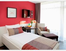 Ein modern eingerichtetes Hotelzimmer mit einer roten Akzentwand, einem Einzelbett mit dekorativen Kissen und einem kleinen Tisch, auf dem Getränke und Obst stehen. Im Hintergrund befinden sich ein Sessel mit Hocker, eine große Fensterfront mit Vorhängen und dekorative Wandbilder.