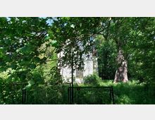 Weißes Gebäude, teilweise verdeckt von dichtem Blattwerk und Bäumen, inmitten einer üppig bewachsenen, grünen Umgebung; im Vordergrund ein kleiner, verrosteter Metallzaun.