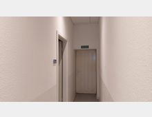 Schmaler, heller Flur mit weiß verputzten Wänden und einer Tür am Ende, auf der ein grünes Schild mit der Aufschrift „Notausgang“ angebracht ist. Links ist eine weitere Tür mit der Beschriftung „K06A“ erkennbar.