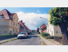 Straßenszene in einer Wohngegend mit mehrstöckigen Häusern im traditionellen Baustil, gekennzeichnet durch rote Ziegeldächer. Am Straßenrand stehen parkende Autos, Gehweg und Grünflächen mit einigen Bäumen säumen die Straße.