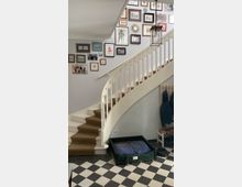 Treppenhaus mit geschwungener, weißer Holztreppe und beigefarbenem Teppichläufer, Wände dekoriert mit zahlreichen gerahmten Bildern. Im Eingangsbereich befinden sich ein Hundebett, an der Wand ein Garderobenbrett mit Jacken sowie Kacheln im schwarz-weißen Schachbrettmuster auf dem Boden.