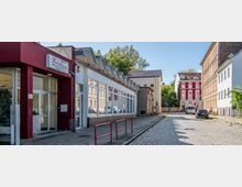 Straßenszene mit Kopfsteinpflaster, links ein modernes Gebäude mit Glasfassade und roter Wand, daneben ein rotes Gebäude mit weißen Fensterrahmen. Im Hintergrund sind Bäume und geparkte Autos zu sehen.