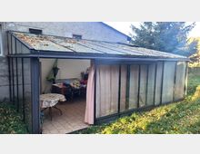 Ein kleiner Wintergarten mit schwarzem Metallrahmen und gläsernen Wänden, teilweise mit Vorhängen bedeckt. Im Inneren befinden sich ein Tisch, Stühle und verschiedene Gegenstände; das Dach ist mit Laub bedeckt.