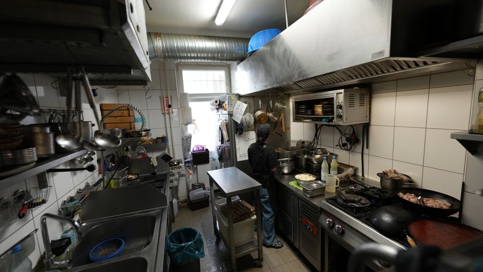 Schmale Großküche mit weißen Fliesenwänden, Edelstahl-Arbeitsflächen und großem Dunstabzug über einem Gasherd mit mehreren Pfannen und Töpfen. Links sind Spüle und Regale mit hängenden Küchenutensilien zu sehen; am Ende des Raums befindet sich ein Fenster und eine Lüftungsleitung unter der Decke.