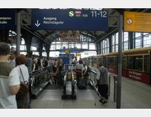 Bahnhofshalle mit Menschen auf Rolltreppen und Bahnsteigen, im Hintergrund ein Schild mit der Aufschrift "Ausgang Reichstagufer". Rechts im Bild steht eine rote S-Bahn, und das Gebäude ist von hohen Fenstern und einer überdachten Stahlkonstruktion geprägt.
