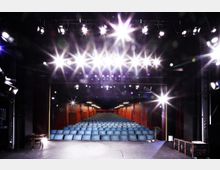 Blick von einer Bühne in einen Theatersaal mit Reihen blauer Sitzplätze, umgeben von roten Wänden. Über der Bühne sind mehrere helle Scheinwerfer angebracht, die den Raum beleuchten.
