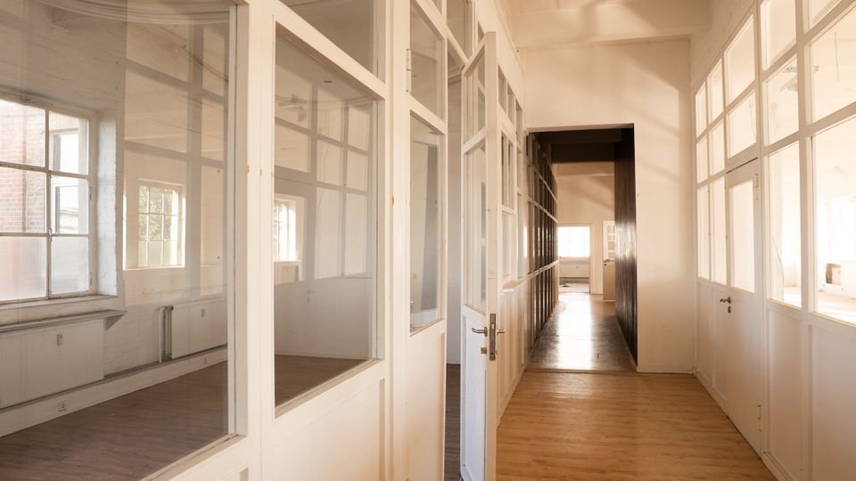 Langer Flur in einem hellen Büro- oder Ateliergebäude mit Holzfußboden und hohen Decken, beidseitig durch weiße Glaswände und Türen in einzelne Räume unterteilt. Große Fenster an den Seiten lassen viel Tageslicht in den Gang.