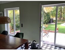 Esszimmer mit großen Fenstern und Blick auf einen Garten. Im Vordergrund ein Holztisch, Stühle und ein geflochtener Deckenlampenschirm, im Hintergrund eine Terrasse mit Gartenmöbeln und grünem Rasen.