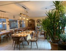 Ein gemütlicher, rustikaler Innenraum mit Holztischen und -stühlen, dekorativem Kronleuchter mit Glühbirnen, Topfpflanzen und hellem Fliesenboden. Im Hintergrund befinden sich ein antiker Schrank, eine Wanduhr und weitere Sitzgelegenheiten.