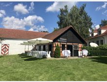 Ein Fachwerkgebäude mit der Aufschrift "Schloss Brennerei" steht in einem gepflegten Garten mit grünem Rasen. Rechts des Gebäudes sind weiße Tische und Stühle unter Sonnenschirmen aufgestellt, umgeben von weiteren Gebäuden mit roten Dächern und weißen Fassaden.