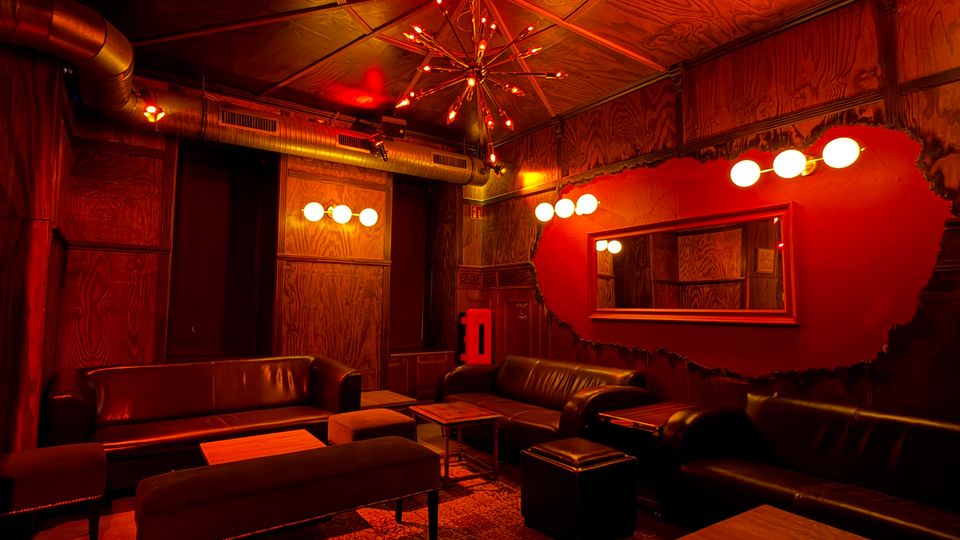 Dunkel beleuchteter Lounge- oder Barraum mit rot-oranger Lichtstimmung, schwarzen Ledersofas und mehreren niedrigen Holztischen. Sichtbare Metall-Lüftungsrohre, eine sternförmige Deckenleuchte und eine große Wandspiegel-Fläche an einer roten Akzentwand prägen den Raum.
