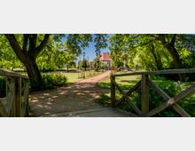 Blick von einer kleinen Holzbrücke auf einen geschwungenen Sandweg durch einen grünen Park mit großen, schattenspendenden Bäumen und Rasenflächen. Im Hintergrund steht ein Gebäude mit rotem Ziegeldach, flankiert von jungen Bäumen und einer Laterne entlang des Weges.