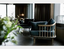 Moderne Lounge mit einem Sessel aus Holz und dunkelblauem Samtpolster, im Hintergrund ein runder Couchtisch, Stühle und große Fenster mit blickdichten Vorhängen. Im Vordergrund unscharfes Grün von Pflanzen.