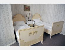 Schlafzimmer mit zwei Einzelbetten im Vintage-Stil, hellen, geschnitzten Holzrahmen und weißen Bettbezügen. Zwischen den Betten steht ein kleiner Nachttisch mit einer Tischlampe, darauf eine Spitzendecke, und an der Wand hängt ein Gemälde mit Blumenmotiv; die Tapete ist dezent gemustert.