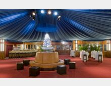 Eleganter Innenraum eines Veranstaltungszeltes mit blauer, drapiert wirkender Decke. Zentral steht ein beleuchtetes Kunstobjekt in einer Schale auf einer Lederbank, umgeben von Hockern, einer Bar mit hohen Sitzplätzen und Stehtischen mit weißen Tischdecken.