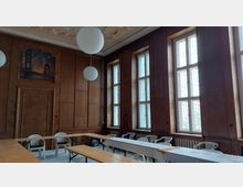 Ein historischer Raum mit holzvertäfelten Wänden, hohen rechteckigen Fenstern mit Gittermustern und einer verzierten Decke. Im Raum stehen Holzbänke und Klapptische mit weißen Tischdecken sowie Plastikstühle, und an einer Wand ist ein Gemälde mit einer Bergbauszene zu sehen.