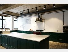 Moderne Küche mit dunkelgrünen Schränken und weißen Arbeitsplatten, einer Kochinsel in der Mitte und einer Wand aus weißen Fliesen. Sichtbetondecke mit schwarzen Scheinwerfern und einem schwarzen Dunstabzug über einem roten Herd.